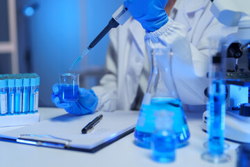 Scientist in gloves and lab coat carefully adding blue liquid to a beaker with a pipette,...