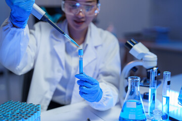 Female scientist in lab coat and protective glasses analyzing blue solution in test tube with...