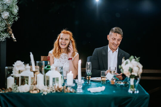  A bride laughs on the phone while sitting beside the groom at a decorated wedding reception table with candles and floral arrangements.