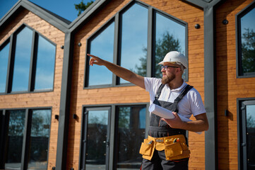 Architect hold plan and pointing on house. Bearded architect wearing hardhat holding project blueprints paper plans standing near comlex of new modern houses looking to something at construction site.