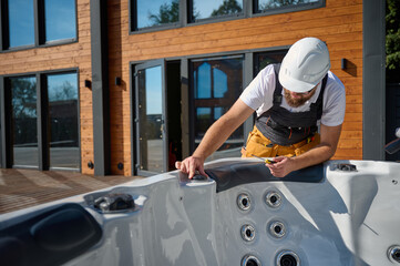 Plumber installing hot tub. Spa technician performing hot tub. Bathtub plumbing. Plumber fixing spa zone. Professional in uniform inspects and prepares a hot tub located on a stylish outdoor patio.