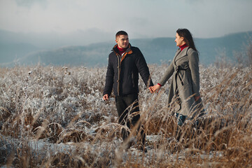 a man and a woman holding hands and walking across a snowy field. The man is wearing a dark jacket and jeans, and the woman is wearing a gray coat and jeans.