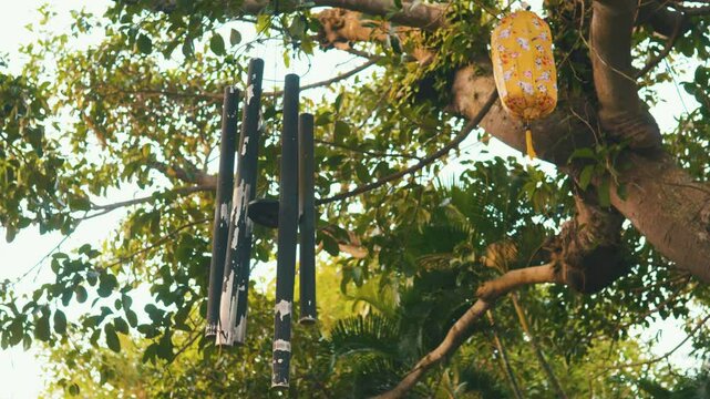 Suspended wind chimes and yellow lantern dangling from tree branch, adding decorative charm to serene garden landscape with soft, peaceful ambiance