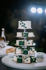 A tiered wedding cake display with white and green mini cakes decorated with leaves, set on a table with elegant tableware..