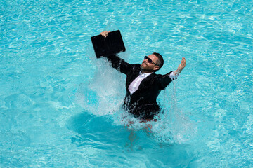Crazy business man in a business suit works for a laptop standing in the water in pool. Remote work. Crazy freelancer. Business and summer. Business man working online with computer laptop in water