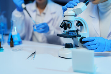 Medical researchers wearing lab coats and gloves are using a microscope and pipette, conducting...