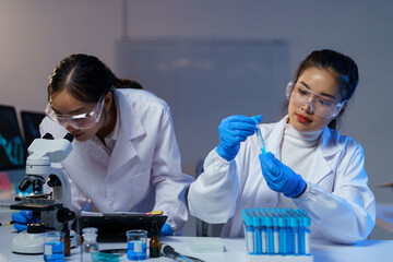 Two female scientists in lab coats and safety glasses conducting research in a modern laboratory,...