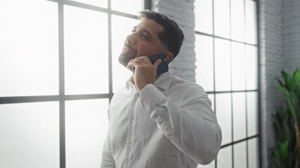 Young man talking on phone in a bright office, smiling and relaxed, with large windows in the background, creating a professional and modern work environment vibe.