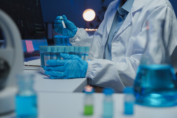 Scientist wearing gloves carefully picking a test tube from a rack in a laboratory at night, with computer monitor and other test tubes in the background, conducting important scientific research