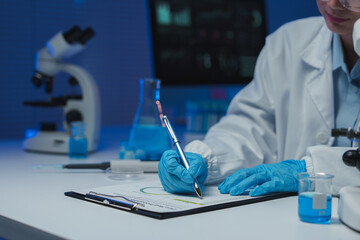 Scientist wearing a lab coat and gloves taking notes on a clipboard in a laboratory setting, with a...