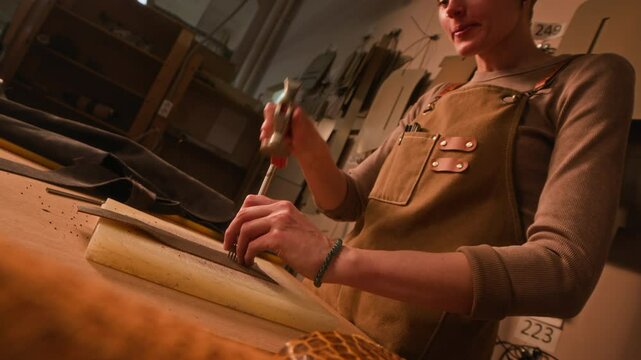 Low angle shot of leather punching process made by professional Caucasian female artisan wearing apron working in small tannery
