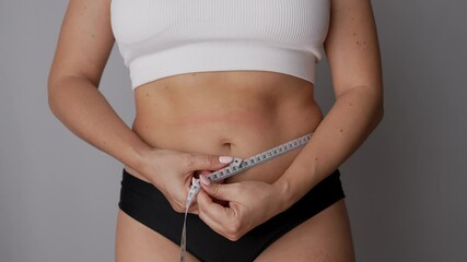 A young woman measuring her belly with a measure tape on a grey background. Diet concept. Overweight, excess weight, weight gain, eating disorder. Belly girth
