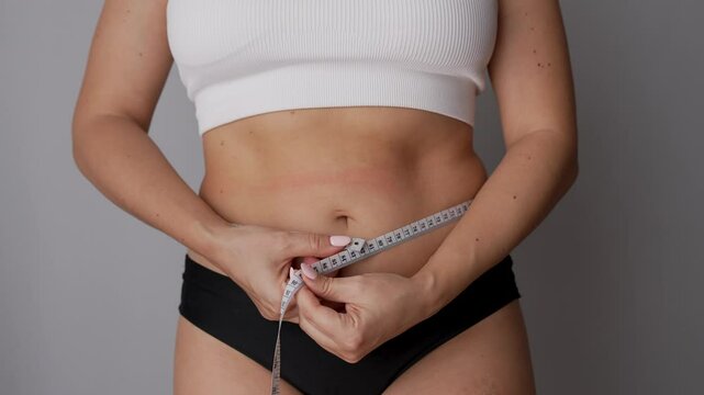 A young woman measuring her belly with a measure tape on a grey background. Diet concept. Overweight, excess weight, weight gain, eating disorder. Belly girth