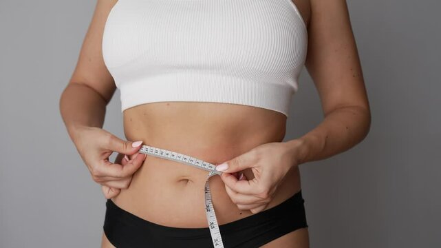 A young woman measuring her waist with a centimeter on a grey background. Diet concept. Overweight, excess weight, eating disorder. Waist girth