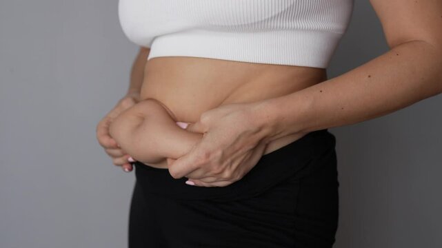 Fat deposits on the abdomen. A young woman holding for belly fat with her hands on a grey background. Overeating, overweight, excess weight, hormonal belly