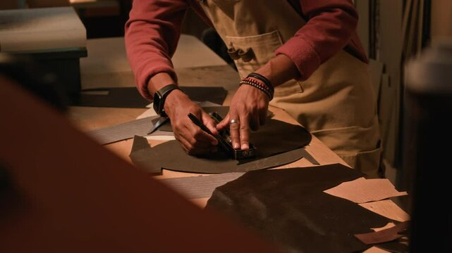 Side view cropped shot of young Black craftsman wearing apron drafting on leather details using special tools, designing handmade bag in equipped tannery workshop with warm lighting