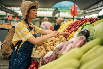 Young asian woman inspects fresh vegetables at outdoor produce stand.