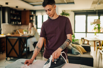 Obraz premium Young woman ironing clothes at home while her partner works on laptop in the background