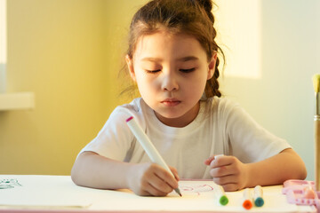 Happy little schoolkid girl drawing in coloring pencils in paper albums, sitting at small wooden studying desk, enjoying creative hobby, kindergarten activity in playroom. Preschool education concept.