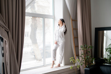 A joyful woman in comfortable pajamas stands by a large window, taking in the peaceful view outside her home during the day