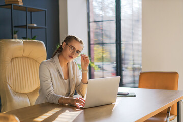 Obraz premium Woman is sitting at a desk with a laptop in front of her. She is wearing glasses and a suit