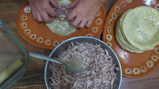 Female hands spooning meat into tortillas to prepare enchiladas. Table with cooking utensils, top view.
