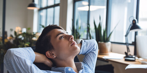 Relaxed Businesswoman in Office Setting. A young woman with short brown hair sits in an office chair, leaning back and gazing out the window with a contemplative expression.