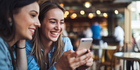 Joyful Friends Sharing Social Media Moment at Cafe. Two young women laughing while looking at smartphone in cozy cafe with warm ambient lighting. 