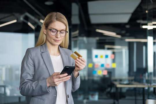 Serious and thoughtful businesswoman with phone and bank credit card in hands. Mature woman at workplace inside office, transferring funds online using banking app, online shopping.