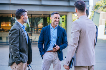 Group of four businessmen in formal and smart casual attire discussing outdoors in a modern urban...
