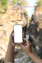 Hands holding a smartphone with white screen in front of a scenic background of rocky cliffs and a flowing river