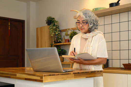 Focused an elderly woman standing at her kitchen counter using her laptop to explore new knowledge
