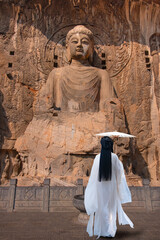 A Chinese woman in traditional white dress holding a traditional white umbrella walks along a stone path - Longmen Grottoes Buddha relief, Luoyang, Henan - View of Longmen Grottoes complex 