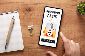 Online scam warning. Overhead shot of hand with smartphone displaying a phishing alert over cozy workspace. 
