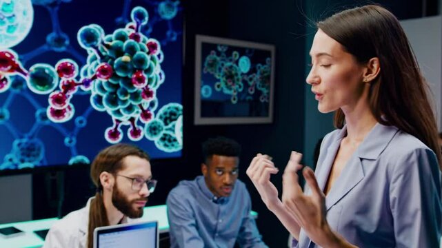 A group of STEM students, both male and female, meet with their project manager in a state-of-the-art lab. A young, female expert is demonstrating a biochemistry presentation on a screen.