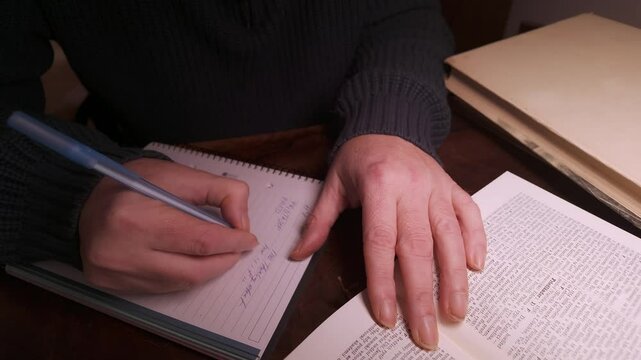 Person is writing notes in a notebook while reading a book at a wooden desk Close-up