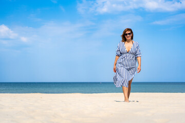 Fototapeta premium Summertime by sea. Beautiful middle-aged woman walking on beach on summer day. Front view. Beauty and self confidence