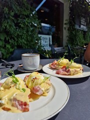 Eggs Benedict with ham, topped with hollandaize sauce and served with coffee at a brunch, outside seating, sunlight table, breakfast