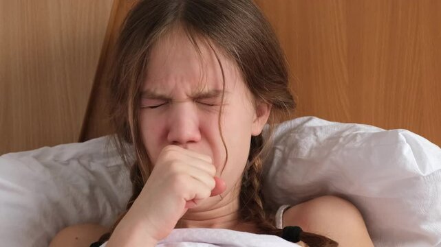 Sick little girl coughing and touching her sore throat with hand, sitting on bed at home, close up. Treatment of illness, flu and cold