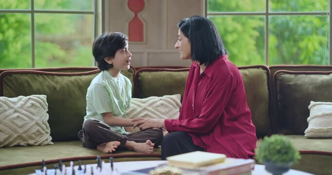 Indian happy adult mother cute kid son sitting on cozy sofa couch talk gossip do tickle have fun joy young lady mom boy child play casual game laugh enjoy good day spend time feel free indoor home