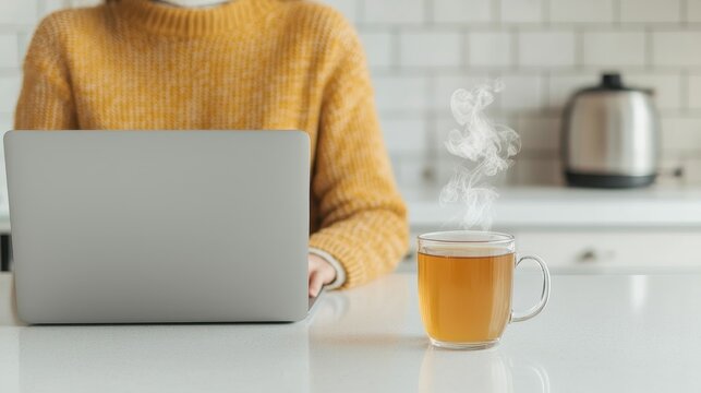 A cozy workspace featuring a person in a yellow sweater, typing on a laptop with a steaming cup of tea beside them.