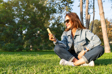 Modern city woman texting on mobile phone while sitting on green lawn in city park.