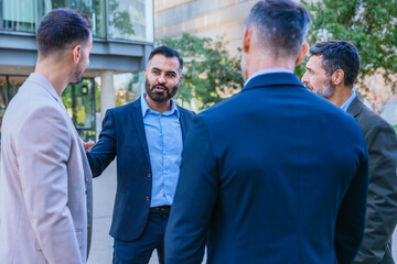 A group of business professionals engaged in a serious discussion outdoors. They are dressed in formal and smart casual attire, with a modern cityscape in the background.