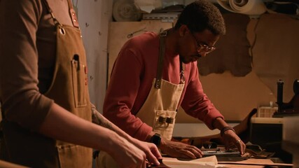 Medium shot of concentrated young Black craftsman applying templates on leather and sharing workbench with unrecognizable colleague in foreground in well-organized craft studio with warm lighting