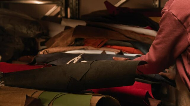 Side view shot of young concentrated Black craftsman in apron standing near store shelves with leather fabric rolls of different colors and choosing one for work