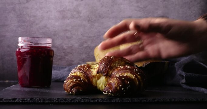 A hand gently places a golden sesame croissant on a slate surface beside a jar of straw or raspberry jam. Rustic bread rolls and a soft blue cloth add charm to the background in warm, natural lighting