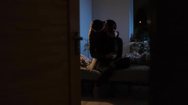 A young couple sits on a bed, hugging and kissing under warm evening lighting, sharing love and affection. French kiss