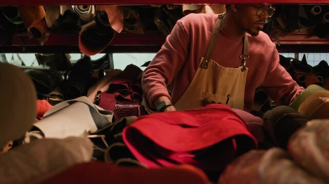 Medium shot of young African American artisan in apron looking for leather textile among multiple leather rolls of different colors storing on shelves in tannery
