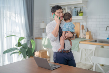 Fototapeta premium Father multitasking by taking a business call while holding his toddler in a bright home kitchen