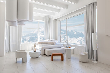 modern pure white interior design of a spacious room features a cozy bed and stylish furniture. Large windows frame a stunning snowy mountain landscape, creating a serene atmosphere during daylight.